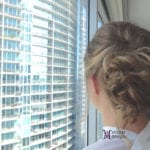A woman with a bun hairstyle looks out an office window with city buildings outside.