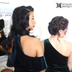 Two women in elegant black dresses with styled hair, facing away.