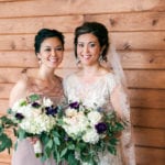 Two women in wedding attire holding floral bouquets, smiling indoors.