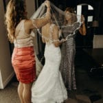 Two women helping a bride adjust her veil indoors.