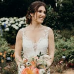 A bride in a white lace wedding dress holding a bouquet of flowers outdoors.