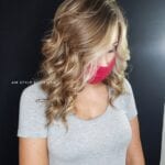 Woman with curly blonde hair wearing a pink face mask and a gray shirt.