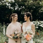 Two women smiling, holding bouquets in a garden.