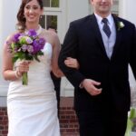Bride in white dress holding purple bouquet, walking with man in suit.