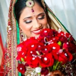A bride smiling while holding a vibrant red flower bouquet.