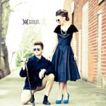 A stylish couple posing outdoors, the woman in a black dress and heels, the man kneeling with sunglasses.