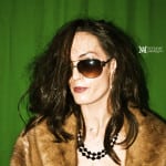 A woman in sunglasses and a fur coat against a green background.