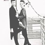 A stylish couple poses on a balcony with a railing.