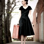 Woman in a vintage black dress holding a suitcase outdoors.