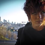 A person with short, curly hair standing outdoors with a city skyline in the background.