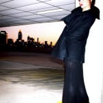 A stylish person in black standing with a city skyline at dusk.