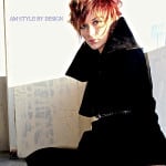 A person with spiky red hair and black clothes sitting indoors.