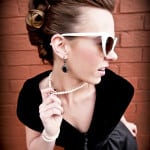 A stylish woman in white sunglasses and pearl necklace poses against a brick wall.