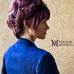 A woman with short, pinkish-purple hair wearing a blue jacket, viewed from the side.