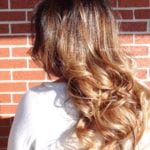 Woman with long, wavy, highlighted hair against a brick wall.
