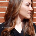 A woman with long brown hair standing against a brick wall.