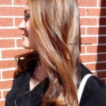 Profile of a woman with long, wavy brown hair against a brick wall.