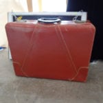 A vintage red suitcase with a black handle on a concrete floor.