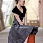 A woman in a black dress sitting on a suitcase outdoors.