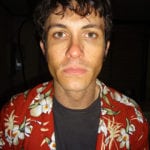 A young man with curly hair wearing a red floral shirt.