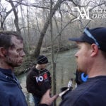 Three men talking outdoors in a wooded area, one with dirt on his face.