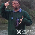 A man in a green sweater with a disheveled appearance near a body of water.