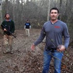 Three men walking on a forest trail, one in the foreground holding a stick.