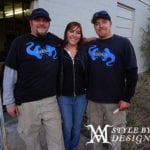 Two men and a woman posing outdoors, men wearing matching black shirts with blue designs.
