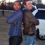 Two men standing back to back outdoors near a white vehicle.