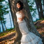 A woman in a white dress leaning against a tree in a forest.