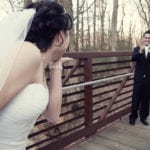 Bride playfully peeking at groom on wooden bridge.