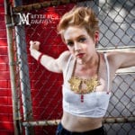 A young woman with tattoos and piercings leans against a chain-link fence.