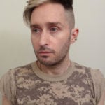 A man with short blonde hair and a camouflage t-shirt looking to the side.