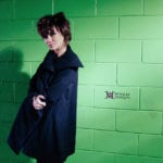 A person in a dark coat standing against a green brick wall.