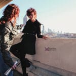 Two women sitting on a ledge overlooking a cityscape on a sunny day.