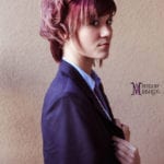 A woman with short pink hair in a dark blazer against a plain wall.