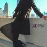A stylish person in black boots and coat posing on a rooftop with a city skyline.