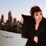 A woman with dramatic makeup posing on a rooftop at sunset with city buildings in the background.