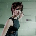 A woman with short hair in a black dress standing against a brick wall.