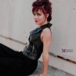 A woman with red hair in a black outfit sits against a wall.