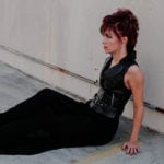 A woman with red hair and black clothing sitting against a wall.