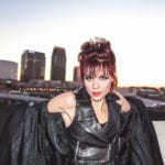 Woman in black leather jacket posing with city skyline at sunset.