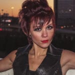 A woman with short auburn hair and striking makeup poses outdoors at dusk.