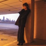 A person in a dark coat leans against a pillar at sunset with a city skyline in the background.