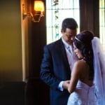 A groom and bride share a tender moment indoors near a window.