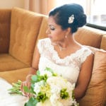 A bride in a white lace dress holding a bouquet, sitting on a brown couch.