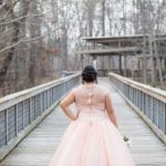 A woman in a pink gown stands on a wooden bridge with her back to the camera.