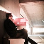 A person sitting against a wall in a parking garage holding a red box.