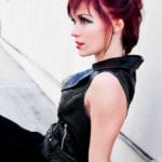 A woman with red hair in a black outfit sitting against a white wall.