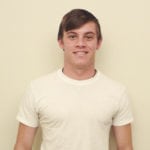 A young man smiling in a plain white t-shirt against a beige background.